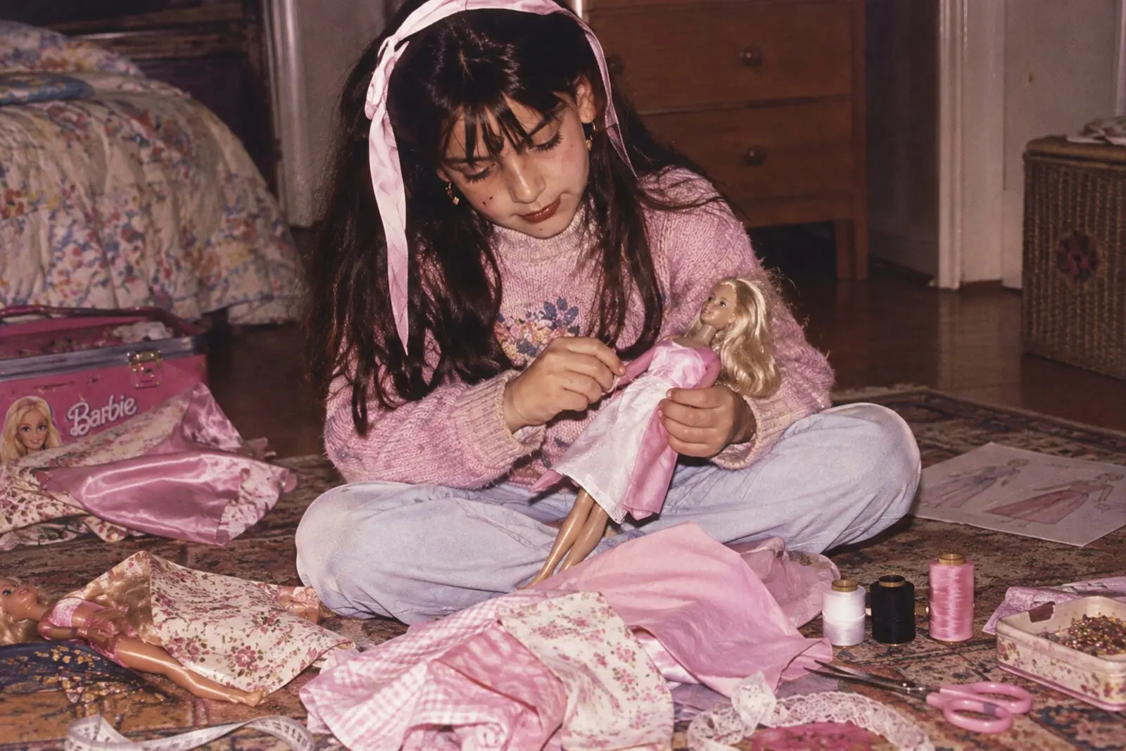 Enfant cousant une tenue pour une poupée Barbie, entourée de chutes de tissu, bobines de fil et ciseaux, illustrant les débuts de la passion pour la couture